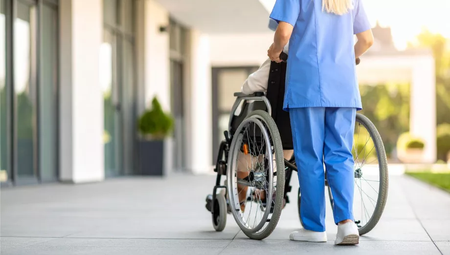 Eine Pflegekraft schiebt einen Patienten im Rollstuhl durch den Außenbereich einer Pflege- oder Gesundheitseinrichtung und unterstützt dessen Mobilität und Betreuung.